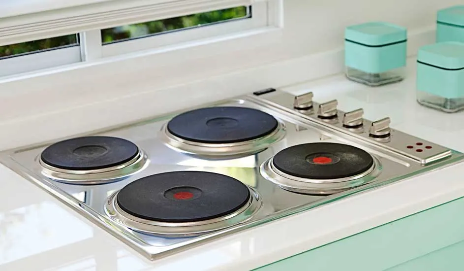 Plaque de cuisson électrique argentée avec quatre brûleurs noirs sur un comptoir blanc, avec des pots turquoise à l'arrière.