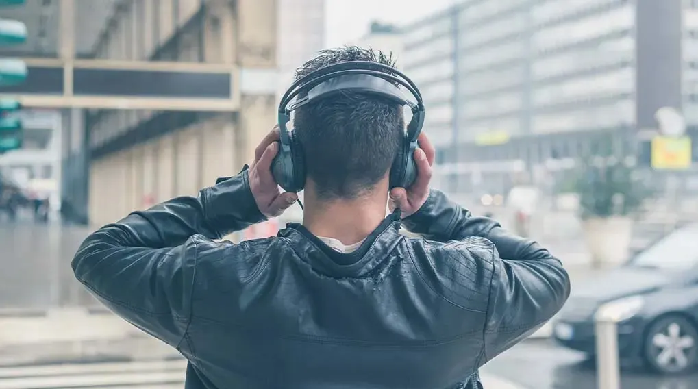 Dos d'une personne avec un casque noir, veste en cuir, debout dans la rue. Bâtiments et voitures en arrière-plan.