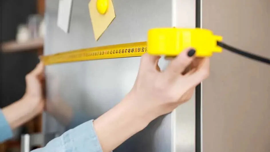 Een geel meetlint wordt tegen een zilverkleurige koelkast gehouden, met gele notities erop.