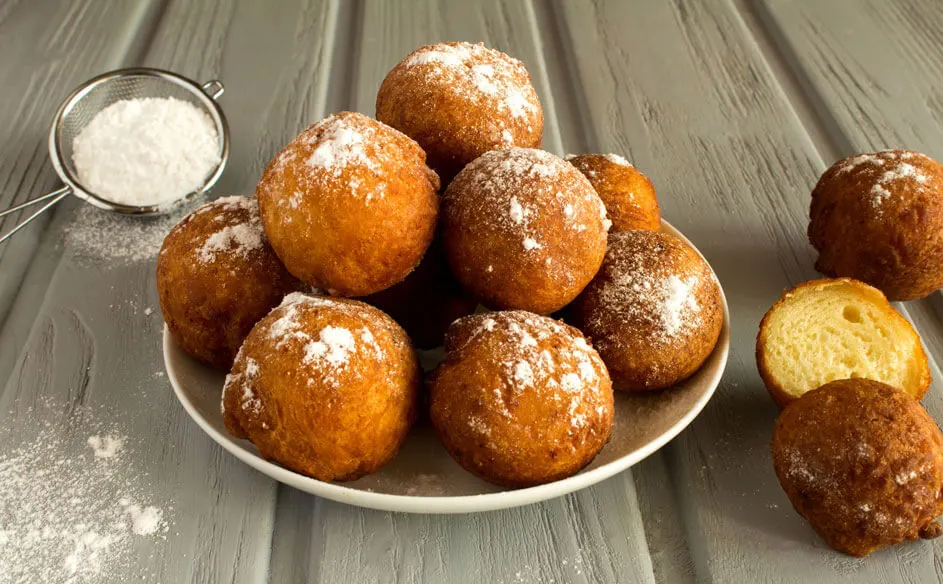 Een stapel gefrituurde oliebollen op een wit bord, bestrooid met poedersuiker. Een zeef met poedersuiker staat ernaast.