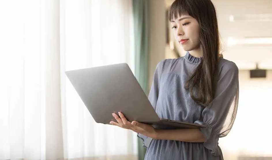 A woman in a light blue blouse holds an Apple MacBook by a window with curtains; Apple logo is visible on the laptop."