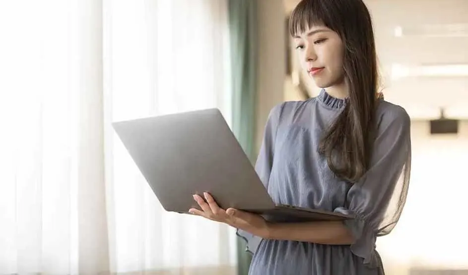 A woman in a light blue blouse holds an Apple MacBook by a window with curtains; Apple logo is visible on the laptop."