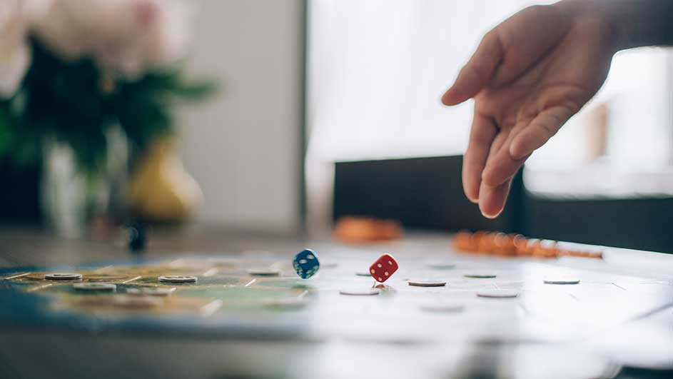 Een blauwe en een rode dobbelsteen op een bordspel, met een hand erboven. Bloemenvaas op de achtergrond.