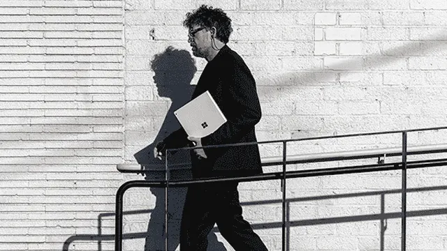 Un homme en costume sombre marche, tenant un ordinateur portable blanc, le long d'une rampe en métal devant un mur de briques blanches.