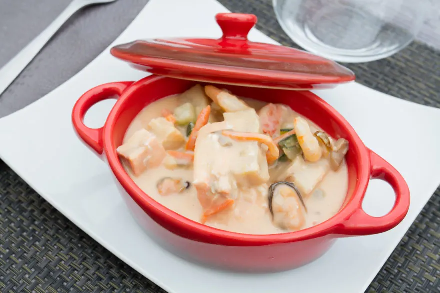 Rode stoofpot met deksel, gevuld met zeevruchten in een romige saus, staat op een wit bord. Een vork en glas zijn zichtbaar op de achtergrond.