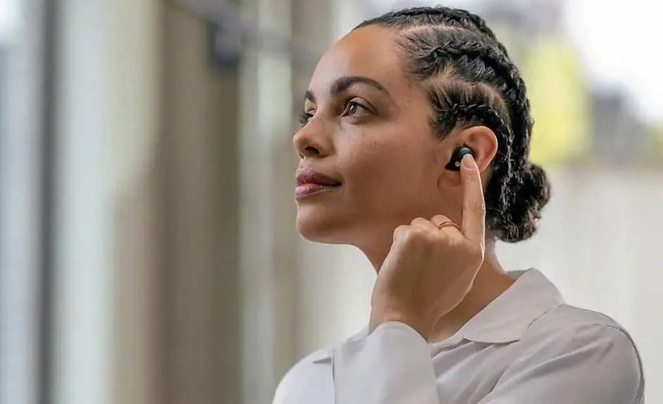 Une personne ajuste un écouteur noir dans son oreille. L'arrière-plan est flou, avec des teintes de blanc et de beige.