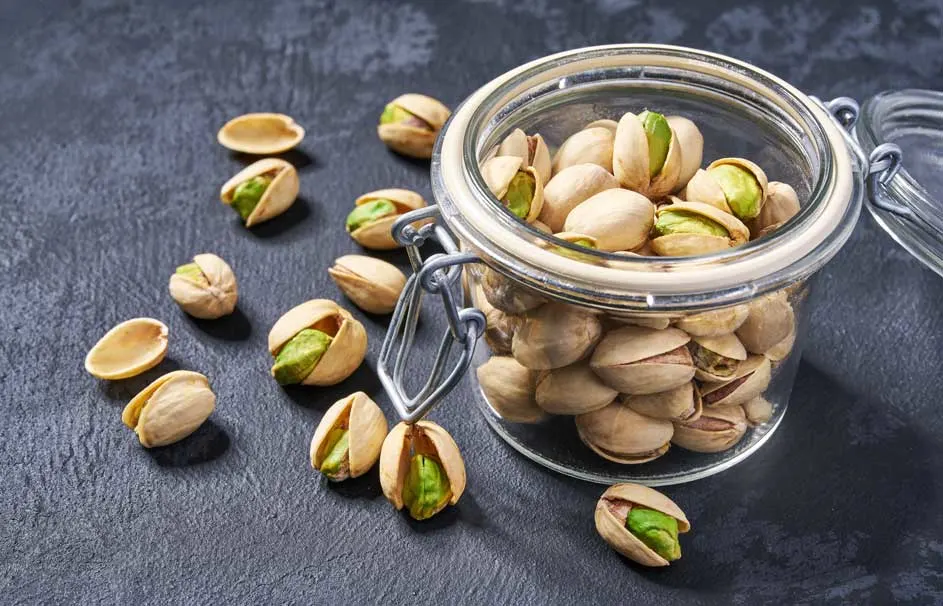 Een glazen pot vol pistachenoten staat op een donker oppervlak, met enkele noten ernaast.
