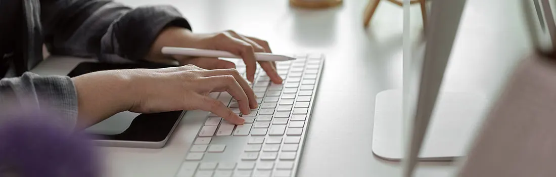 Mains tapant sur un clavier blanc à côté d'un smartphone et d'un stylet sur une table blanche, avec un écran en arrière-plan.