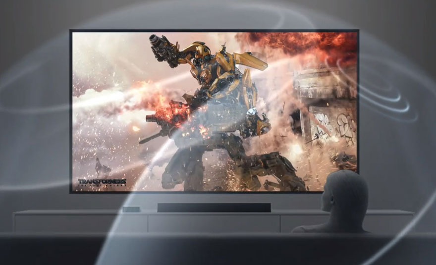 Een grijze tv toont een Transformer in actie, met vuur en rook. De tv staat op een witte kast.