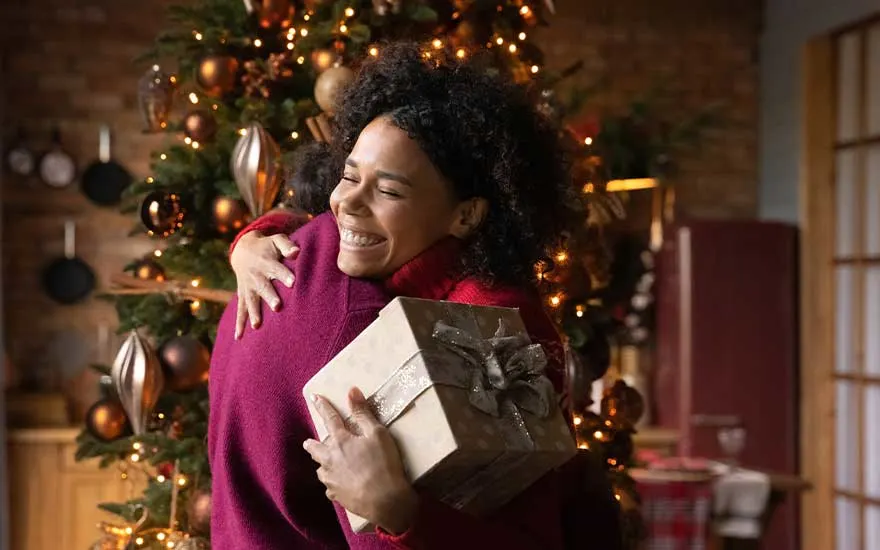Deux personnes s’enlacent devant un sapin illuminé, tandis que la femme souriante tient un cadeau soigneusement emballé dans ses mains.