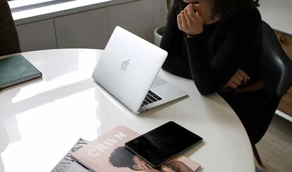 Une personne est assise à une table avec un MacBook Apple ouvert, magazines et iPad à côté ; le logo Apple est bien visible.