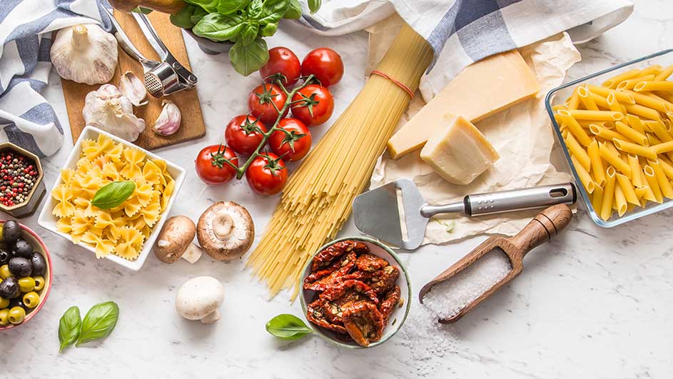 Diverse ingrediënten voor pasta, zoals spaghetti, penne, tomaten, kaas, knoflook en basilicum, op een witte marmeren ondergrond.