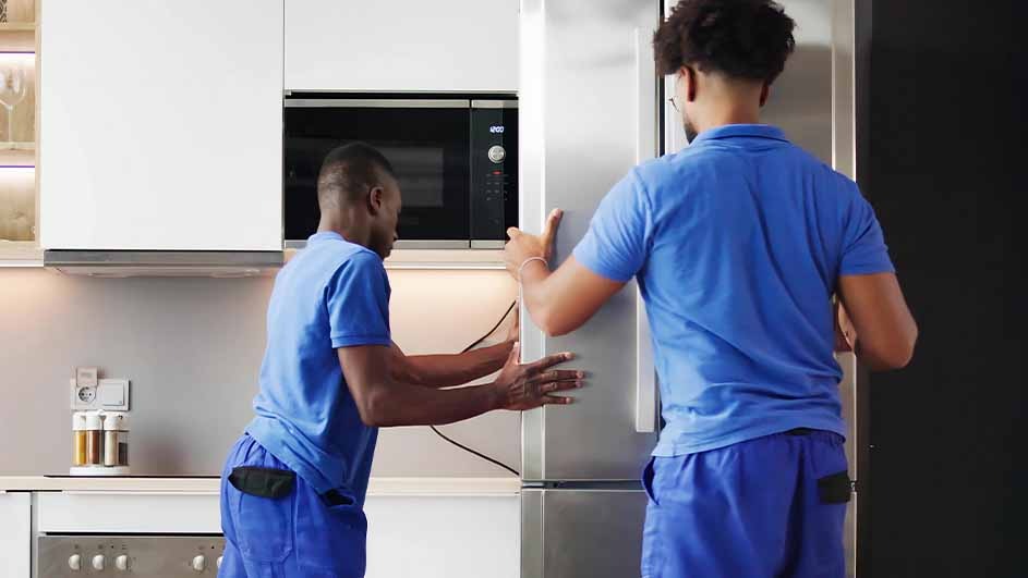 Two workers in blue uniforms install a stainless steel refrigerator in a modern kitchen with white cabinets and a built-in microwave.