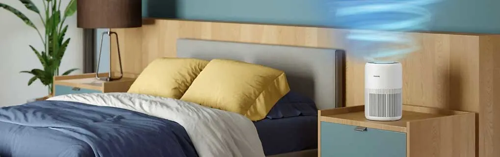 Slaapkamer met houten bed en nachtkastjes; een witte luchtreiniger blaast blauwe wervels boven het kussen in rustige, opgeruimde ruimte.