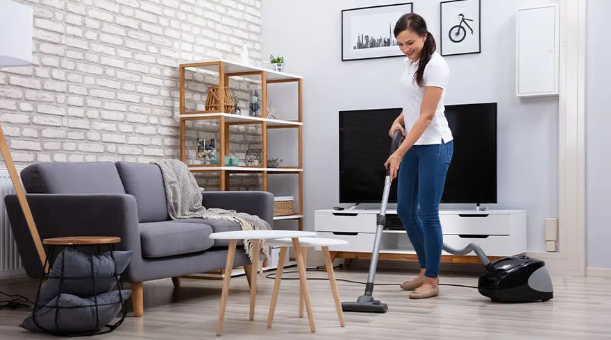 A living room with a grey sofa, wooden shelves, and a TV. A black vacuum cleaner is on the floor.