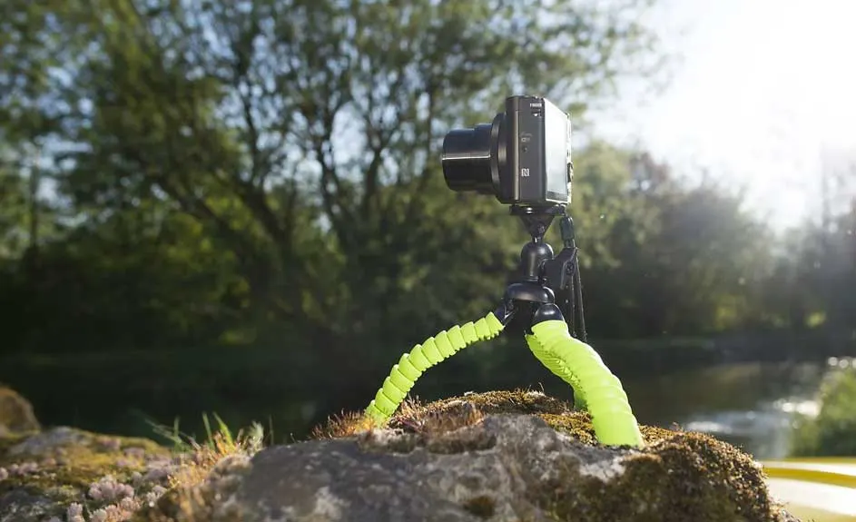 Een camera op een groen statief staat op een rots met een achtergrond van bomen en water.
