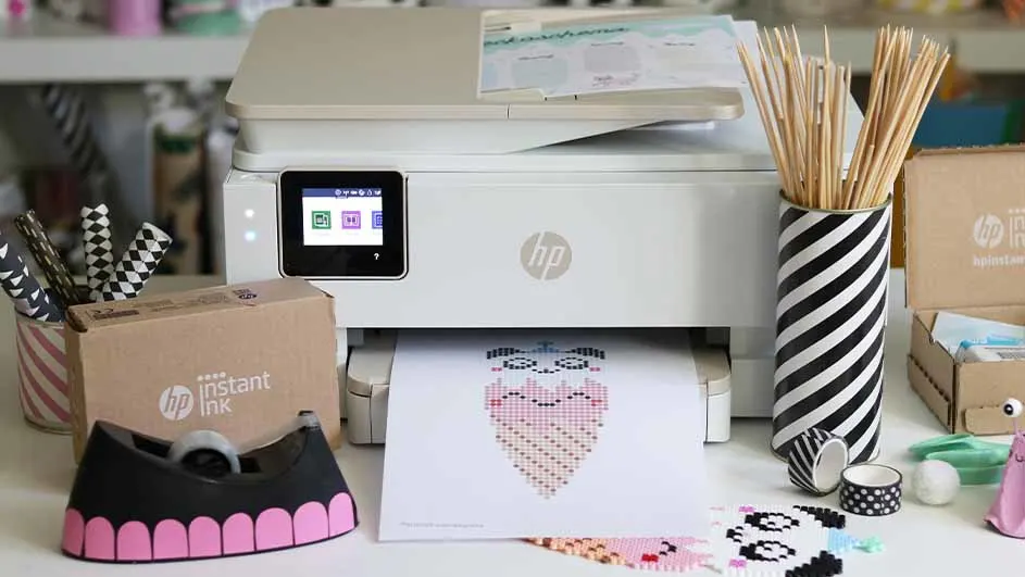 Een witte HP-printer print een pixelhart. Er staan pennen, een doos, en een zwart-wit gestreepte vaas op de tafel.