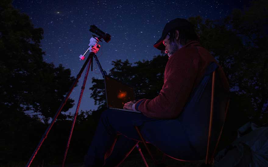 Sous un ciel nocturne rempli d’étoiles, une personne utilise un ordinateur près d’un télescope, probablement pour observer le ciel.