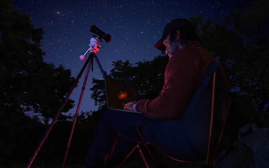 Sous un ciel nocturne rempli d’étoiles, une personne utilise un ordinateur près d’un télescope, probablement pour observer le ciel.