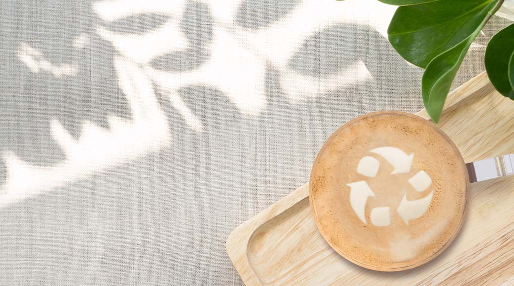 Een cappuccino met een recyclesymbool op een houten dienblad, naast groene bladeren en een lichte achtergrond.