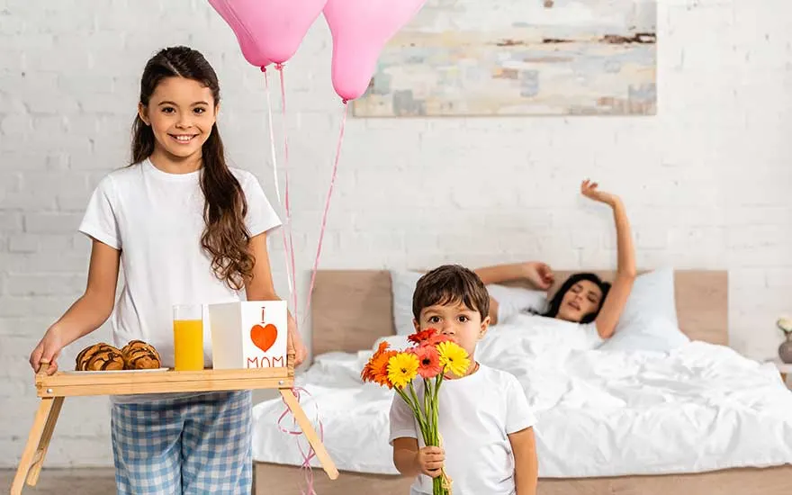 Twee kinderen brengen ontbijt op bed. Een meisje draagt een dienblad met gebak, sap en een kaart. Een jongen heeft bloemen.