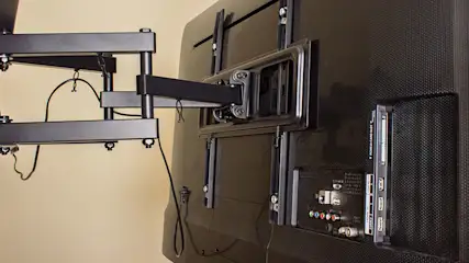 Een zwarte tv is met een metalen beugel aan een beige muur bevestigd, kabels hangen los.