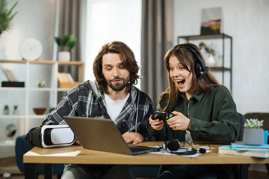 Two people sit at a table with a laptop; the woman in a headset cheers, holding a controller, and a VR headset is lying nearby too.
