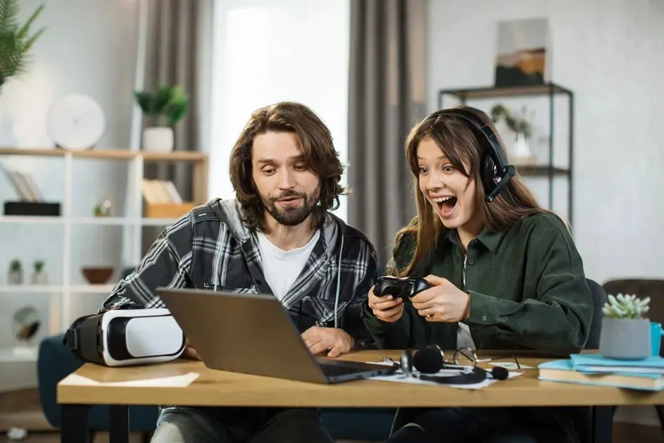 Two people sit at a table with a laptop; the woman in a headset cheers, holding a controller, and a VR headset is lying nearby too.