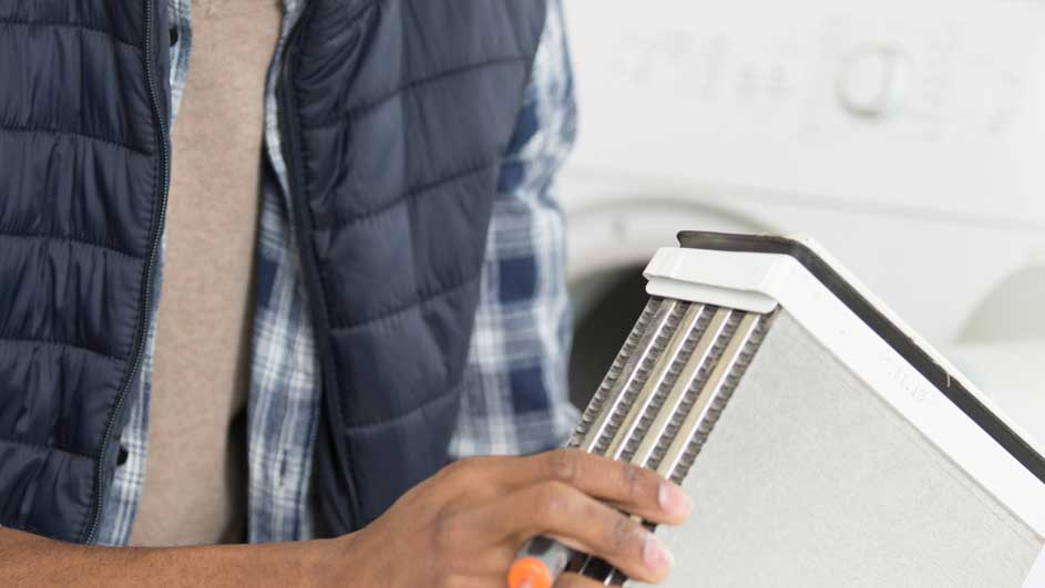 Een man in een blauw vest en geruit overhemd houdt een wit luchtfilter vast voor een wasmachine.