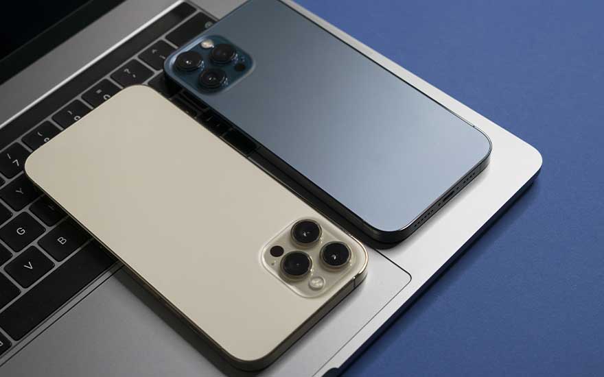 Deux iPhone modernes, l’un doré et l’autre bleu, sont posés côte à côte sur le clavier d’un ordinateur portable, sur un fond bleu uni.
