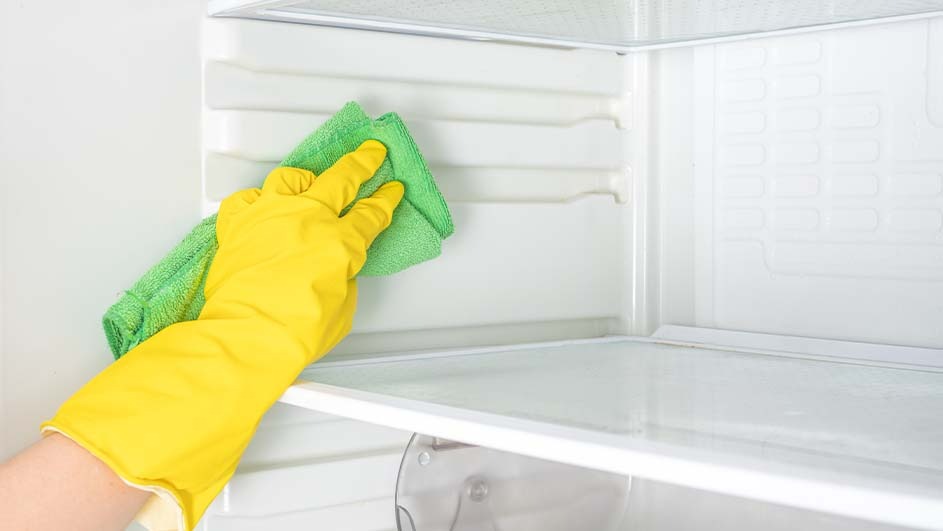 A gloved hand cleans a white refrigerator interior with a green cloth. Shelves are visible in the background.