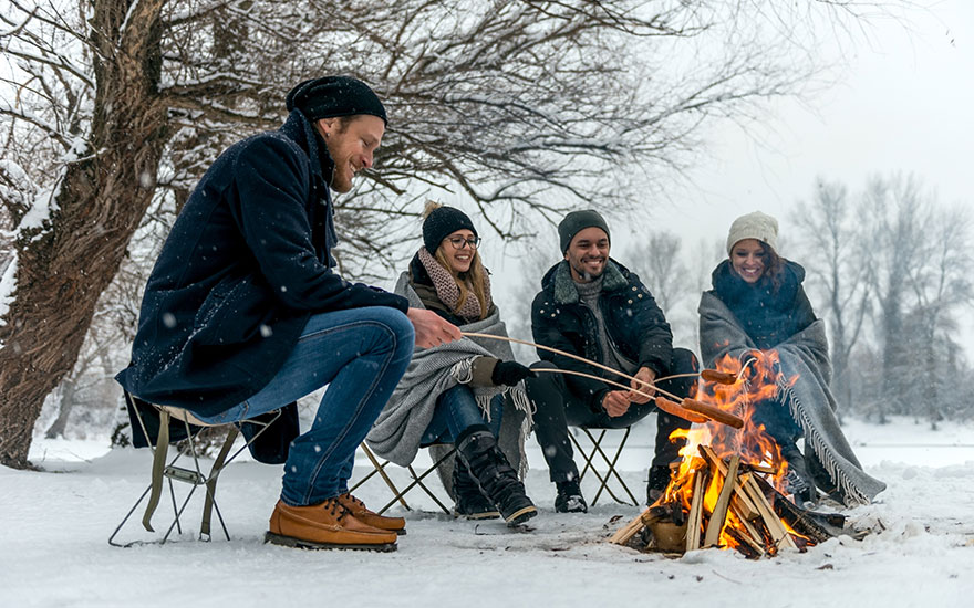 Vier mensen zitten op klapstoelen rond een vuurkorf in de sneeuw. Ze roosteren worstjes aan stokken. Achtergrond: bomen en sneeuw.