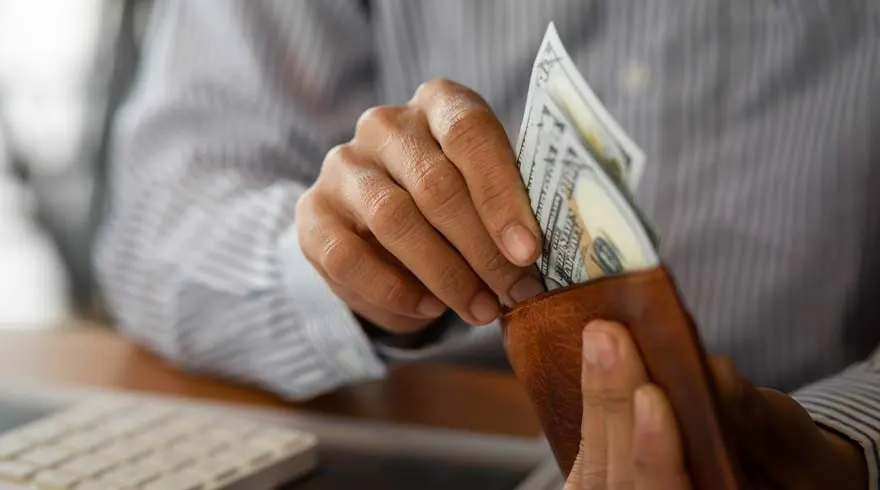 Handen stoppen dollarbiljetten in een bruine portefeuille. Een toetsenbord is zichtbaar op de achtergrond.