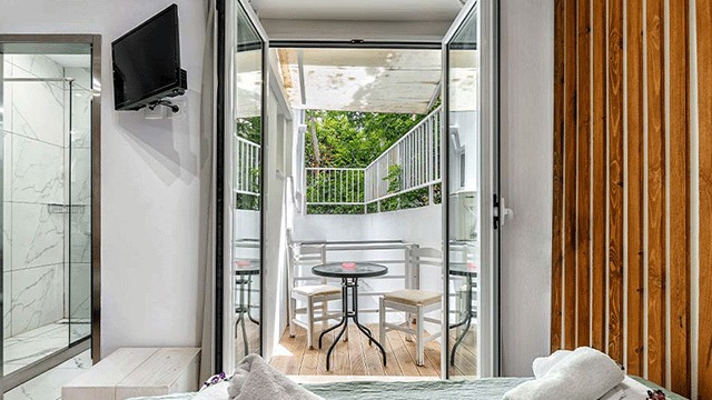 Chambre avec TV, douche et balcon. Balcon avec table et chaises, vue sur la verdure. Panneau mural en bois.