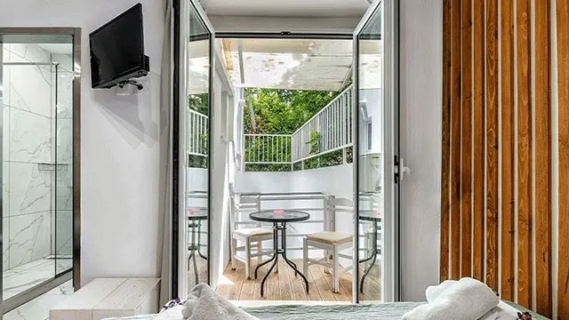 Chambre avec TV, douche et balcon. Balcon avec table et chaises, vue sur la verdure. Panneau mural en bois.