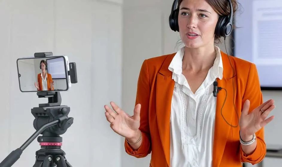 Een vrouw in een oranje blazer en koptelefoon spreekt naar een smartphone op een statief. Ze gebaart expressief terwijl ze in een studio tegen de camera praat. Op het scherm is haar videoafbeelding zichtbaar.