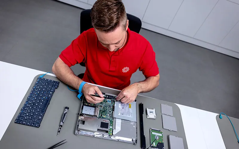Medewerker in een rood poloshirt repareert een geopende laptop aan een werktafel met gereedschap en onderdelen.