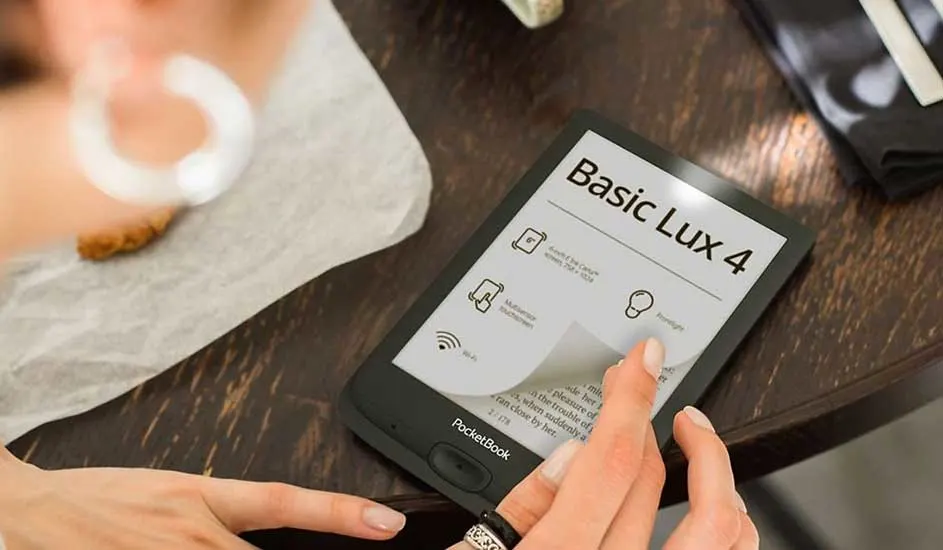 E-reader "Basic Lux 4" op houten tafel, met hand. Scherm toont menu met pictogrammen. Achtergrond: doek, koekje en tas.
