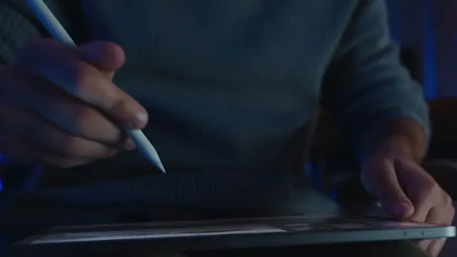 Un homme utilise un stylet blanc sur une tablette grise. L'arrière-plan est sombre avec un éclairage subtil.
