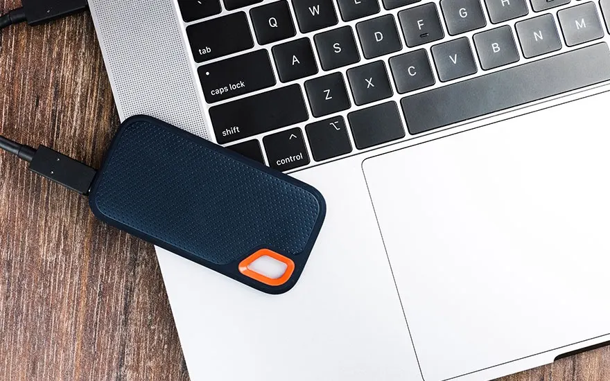 Disque dur externe bleu connecté à un ordinateur portable argenté sur une table en bois. Le clavier est noir et gris.