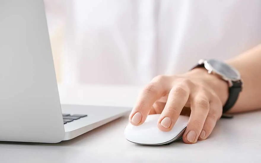 A person wearing a watch uses a white wireless mouse next to an open laptop on a bright, tidy desk.