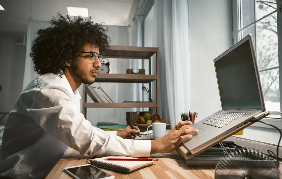 Un bureau avec un ordinateur portable ouvert, un bloc-notes, un téléphone et une tasse blanche devant une fenêtre.