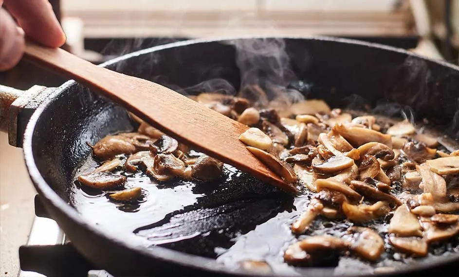 Une poêle noire remplie de champignons bruns tranchés est en train de cuire sur une cuisinière. Une spatule en bois remue les champignons.