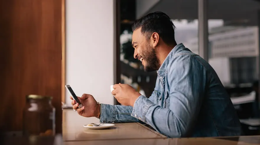 Een man zit aan een tafel met een telefoon en een kopje, gekleed in een blauw overhemd. De achtergrond is een wazig interieur.