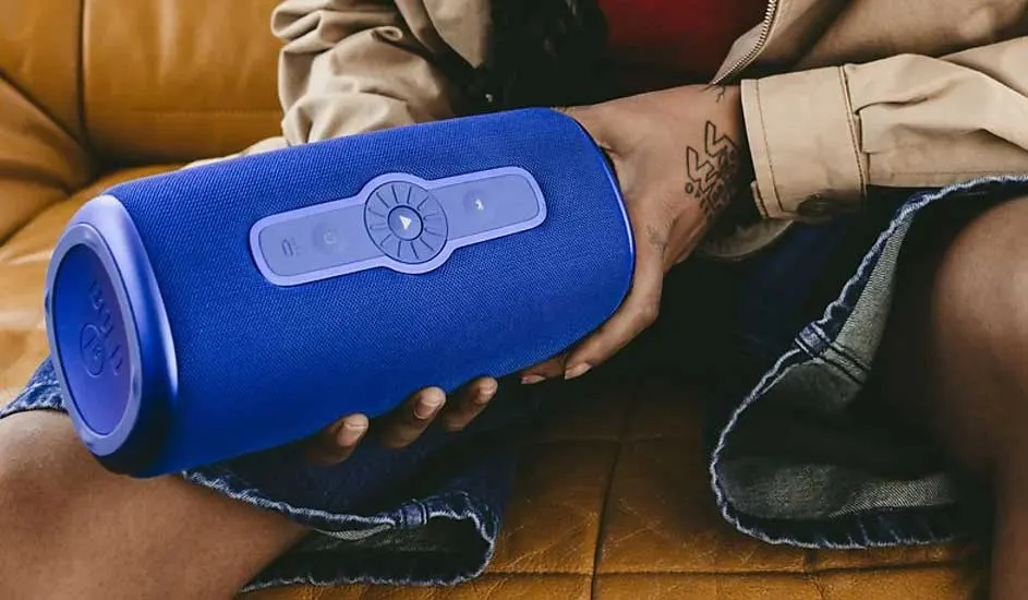 Voici une enceinte Bluetooth JBL bleue tenue sur les genoux de quelqu’un assis sur un canapé jaune, avec des boutons de commande visibles dessus.