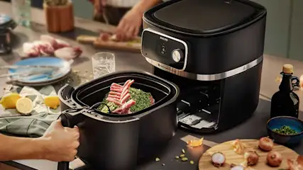 Un airfryer noir près d'une main tenant un panier avec de la viande et des herbes. Des citrons et des oignons sont visibles.