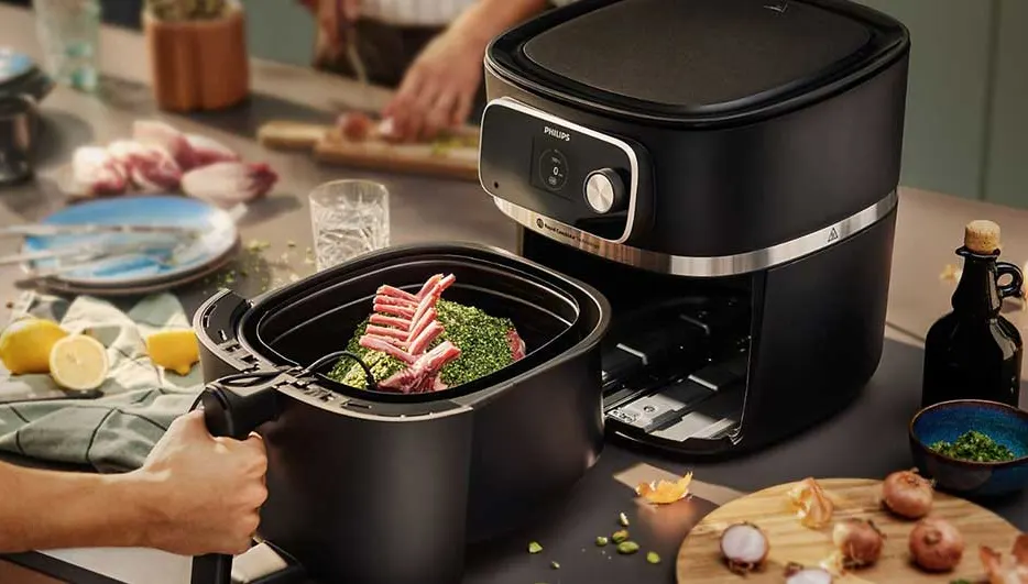 Un airfryer noir près d'une main tenant un panier avec de la viande et des herbes. Des citrons et des oignons sont visibles.
