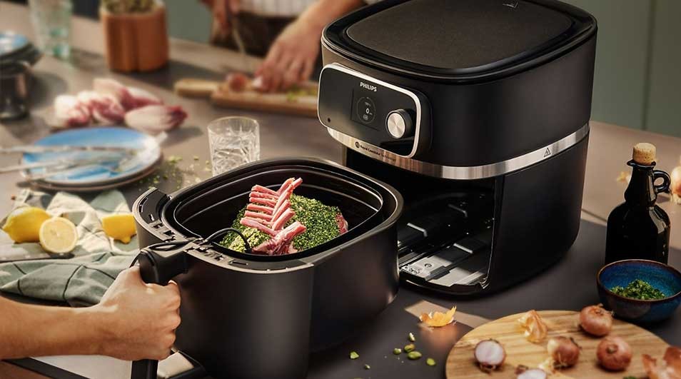 A black air fryer sits on a counter next to raw meat in a basket. Lemons and plates are in the background.