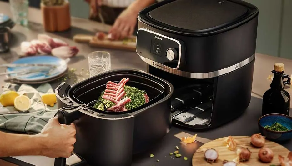 A black air fryer sits on a counter next to raw meat in a basket. Lemons and plates are in the background.
