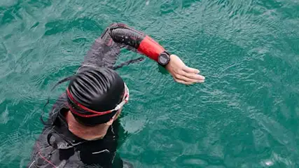 Een zwemmer in zwart wetsuit en badmuts zwemt in groen water, met een sporthorloge om de pols.